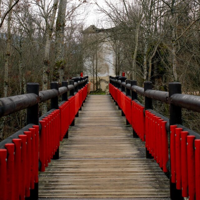 Puente de madera con barandal rojo y postes oscuros, perspectiva centrada hacia el final. Al fondo asoma una pequeña construcción entre árboles desnudos. El rojo aquí no grita: ordena el paso.