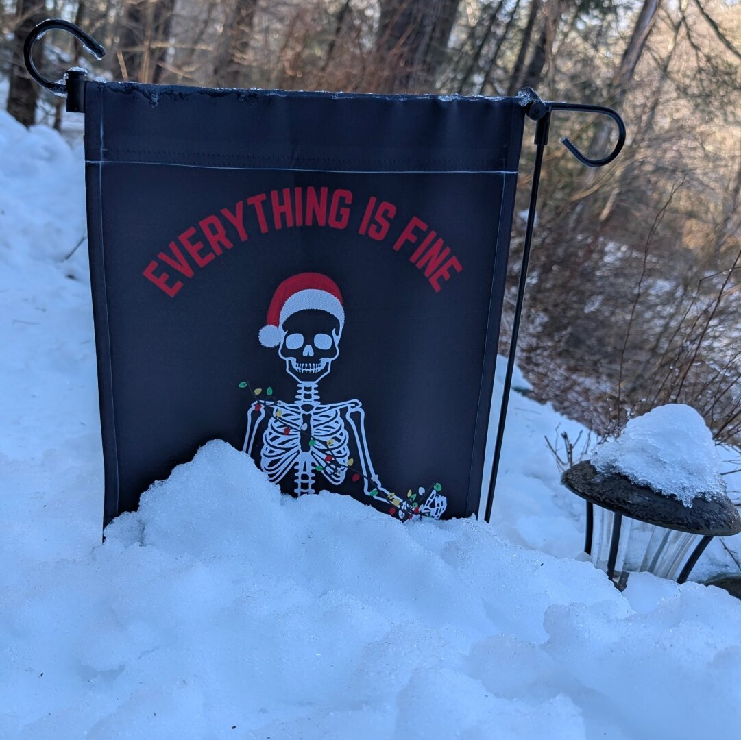 A garden flag depicting a cross legged skeleton in zen pose, draped in colored lights and a santa hat with the words "EVERYTHING IS FINE" over its head. The flag is half buried in snow with thick ice over the top