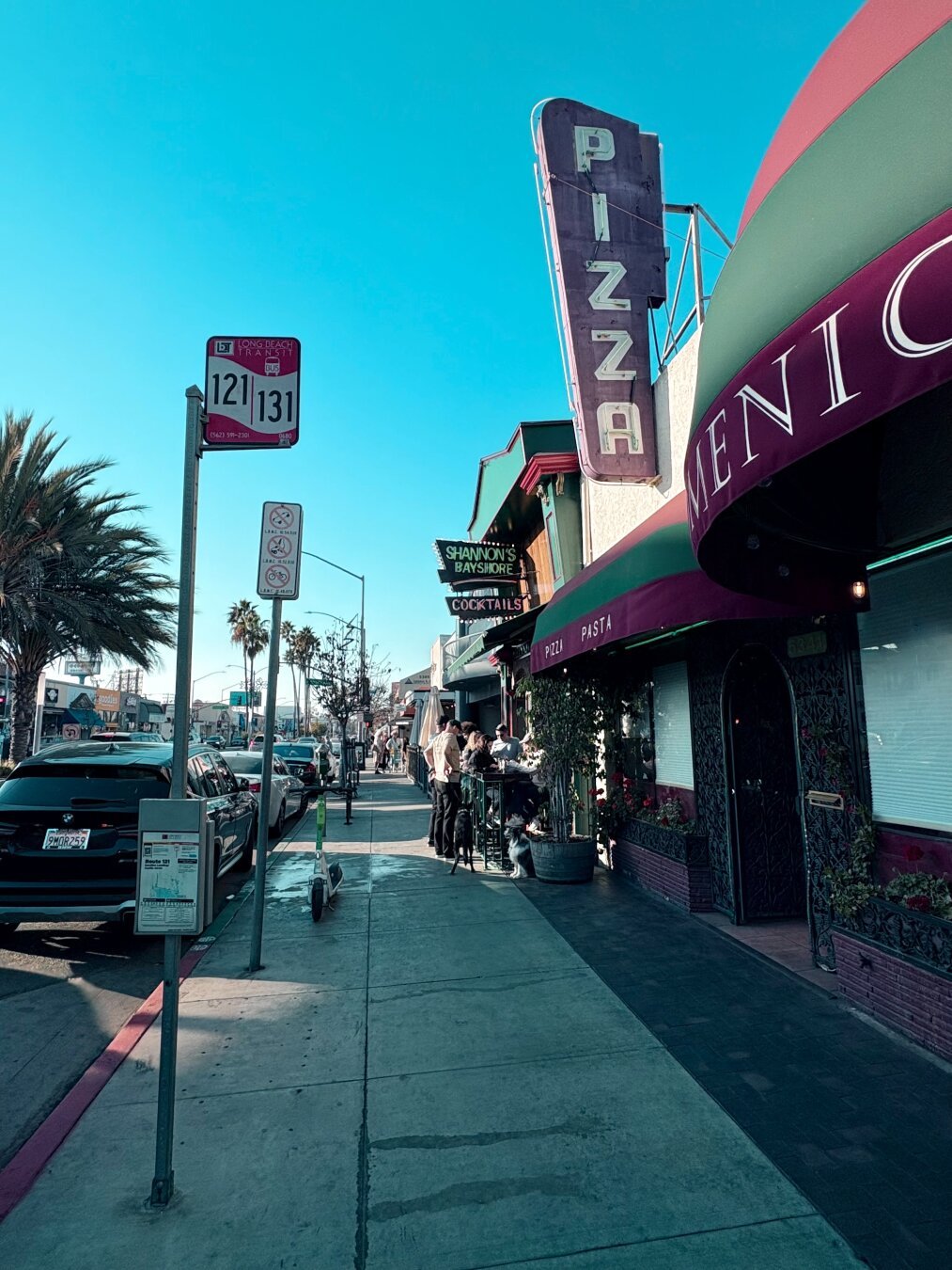 View from the sidewalk, with parked cars and the street in the left, restaurants with outdoor seating on the right. There are several people eating outside, and a couple dogs.