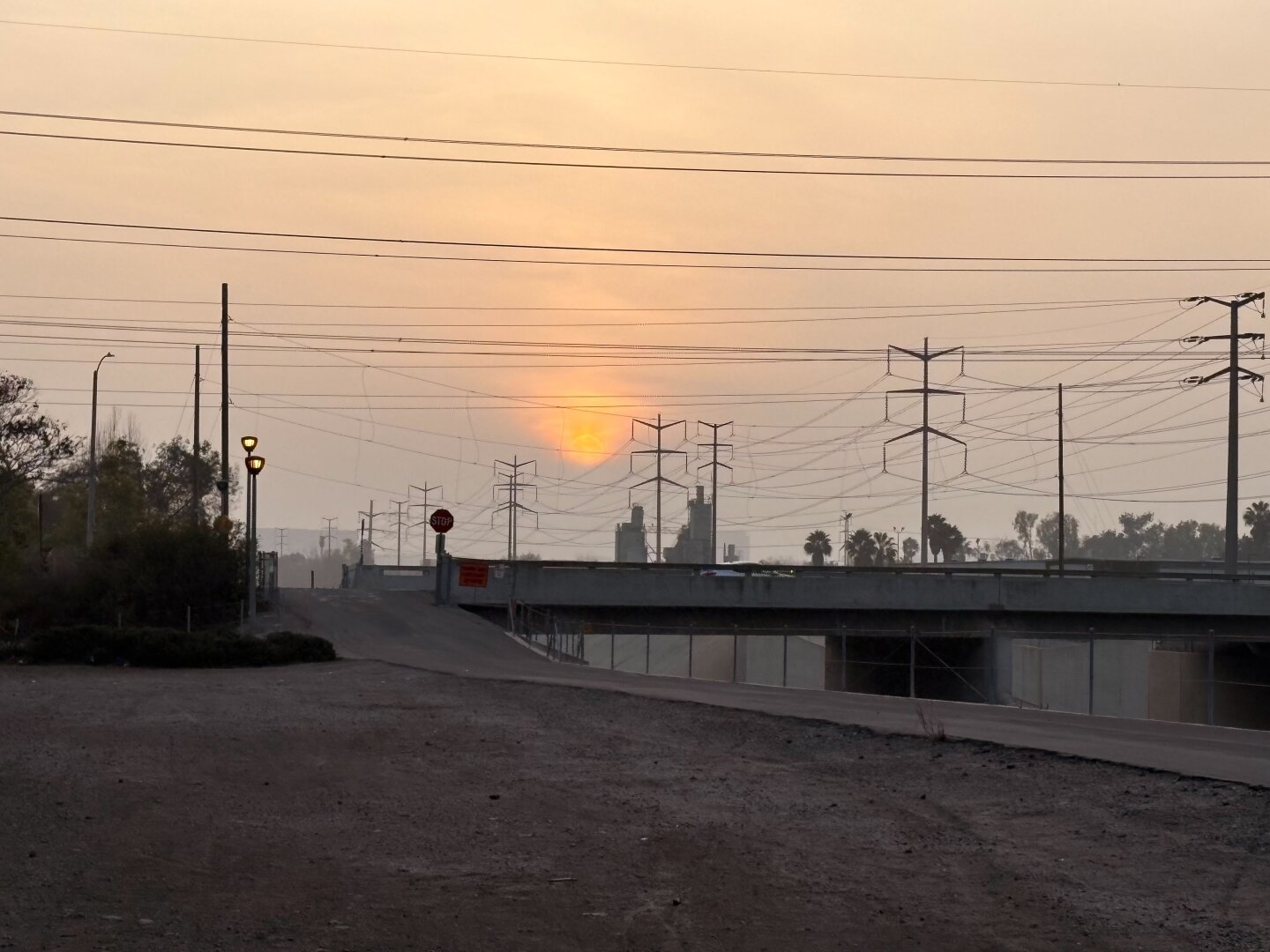 The setting sun over a bridge over a channel in California. Power lines crisscross the foreground. The street lights are just coming on, and the sky is hazy. Shot on iPhone 16.