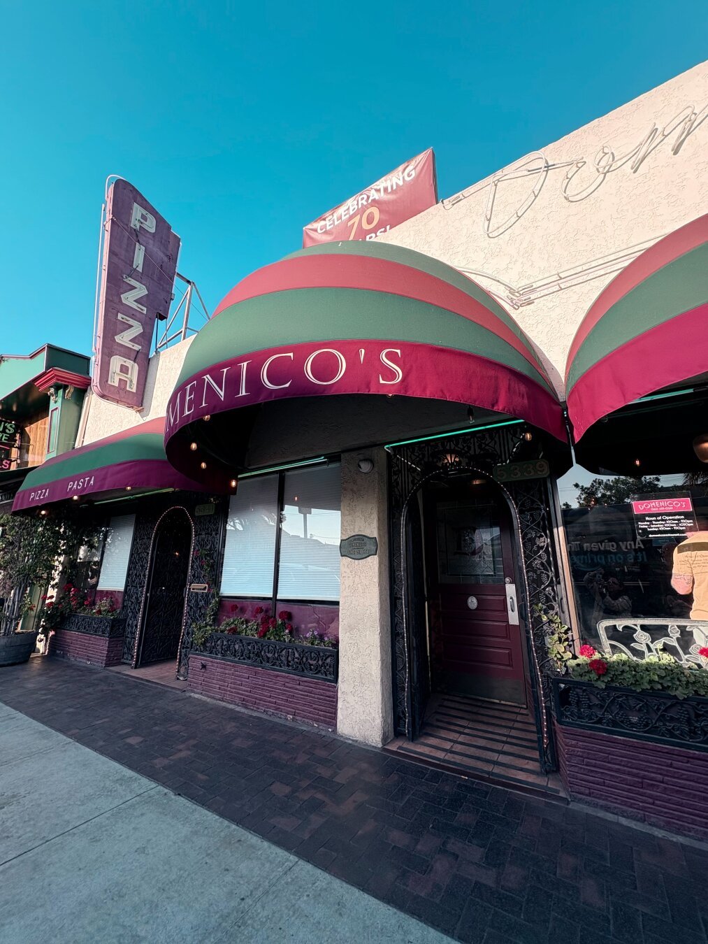 An Italian-inspired storefront with large green and red canopy over the door reading “Domenico’s”. Above the canopy to the left is a large neon sign reading “Pizza”, vertically. It’s mid-afternoon so the light is soft with lots of shadows, and the neon sign is not yet lit.