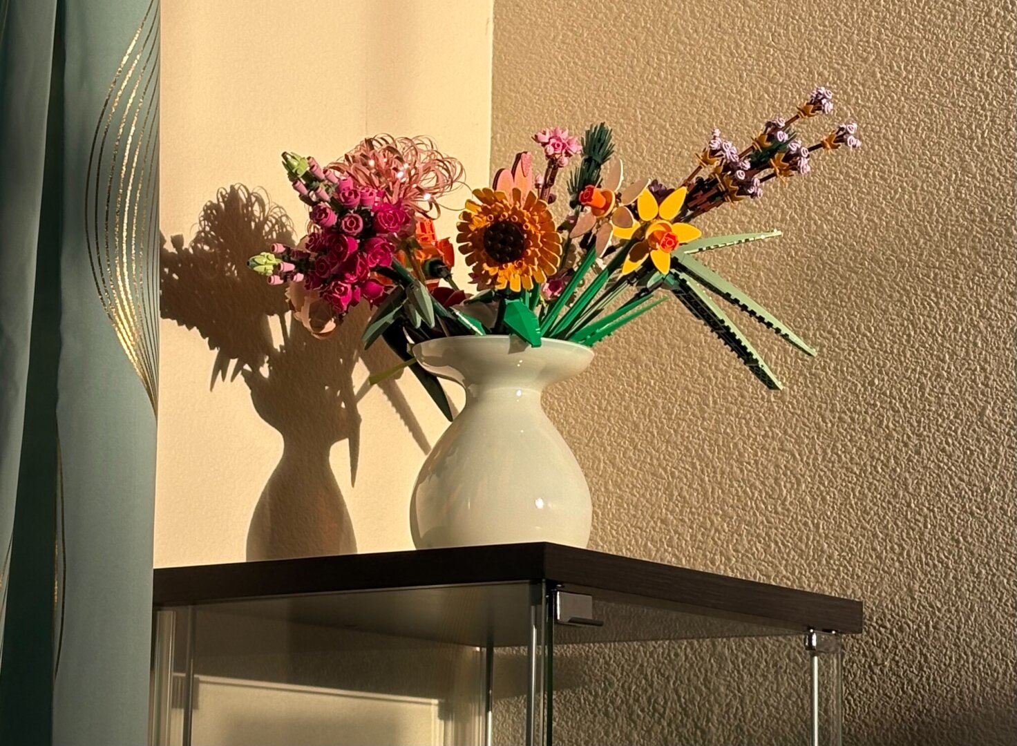 A milky green vase atop a glass display cabinet, full of Lego flowers. Lit by warm, rosy morning sun.