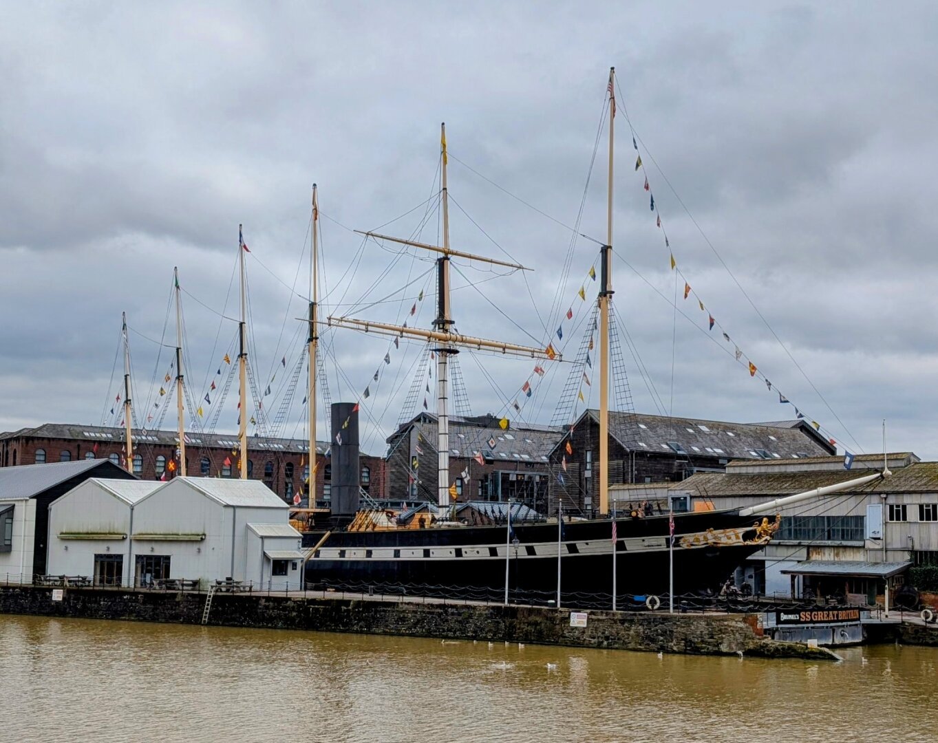 image:A large black steamship, the SS Great Britain, docked at a pier. The ship has several tall masts with colorful flags hanging between them. Behind the ship are industrial-style brick buildings and a more modern white warehouse building next to it. The water in the river is brown and looks somewhat murky. The sky is overcast.