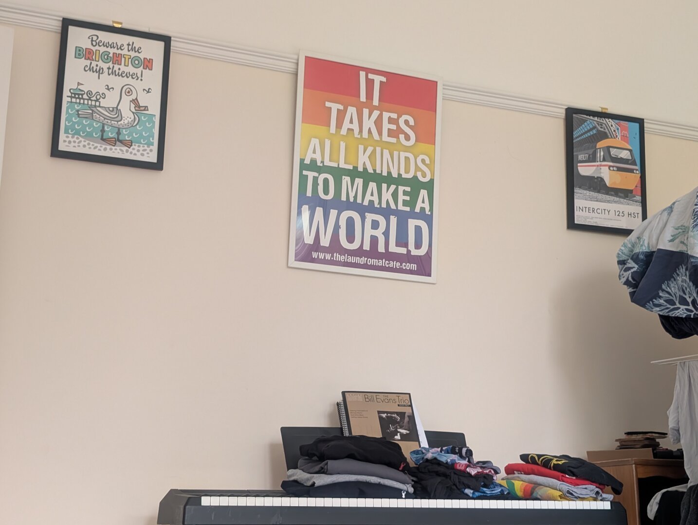 A shot of an off-white wall, with three posters. A larger one in the middle that says "It takes all kinds to make a world", with rainbow stripes in the background. Flanking it are smaller posters, one cartoon of a seagull with a fry in its mouth and the words "Beware the Brighton chip thieves", the other a stylized drawing of an Intercity 125 HST in Intercity Swallow colours.
