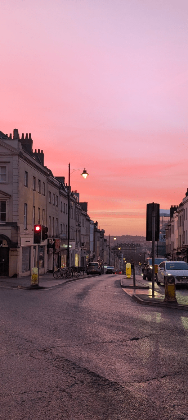 A picture of the top of a street surrounded by buildings at sunrise, with pink hues fading into red, then yellow, on the horizon.