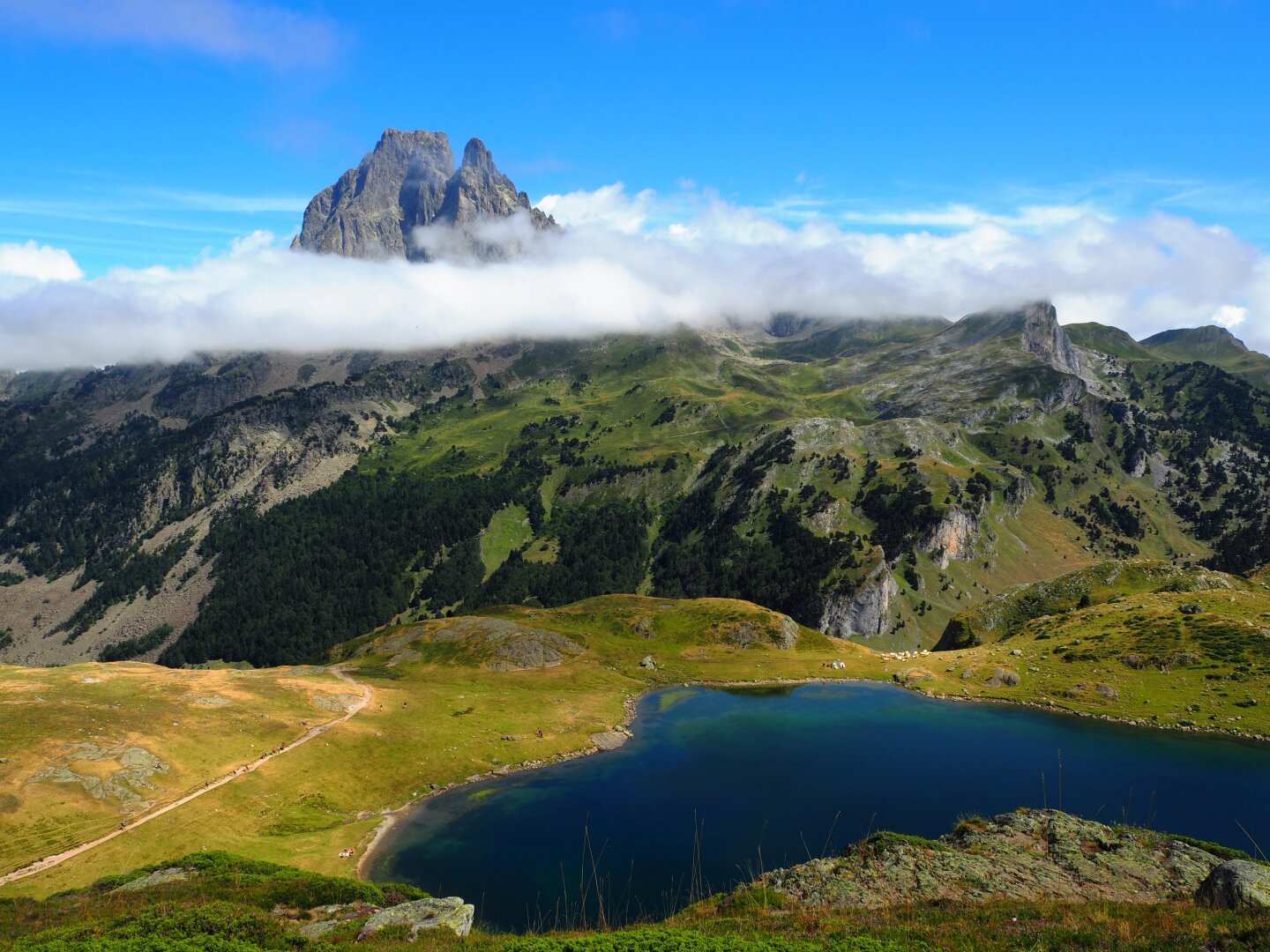 Photo du Pic du midi d’Ossau au-dessus d’une bande de nuage en arrière plan. Au second plan, la vallée le sépare du lac Roumassot au premier plan. Le ciel est bleu, le lac est en forme de cœur.