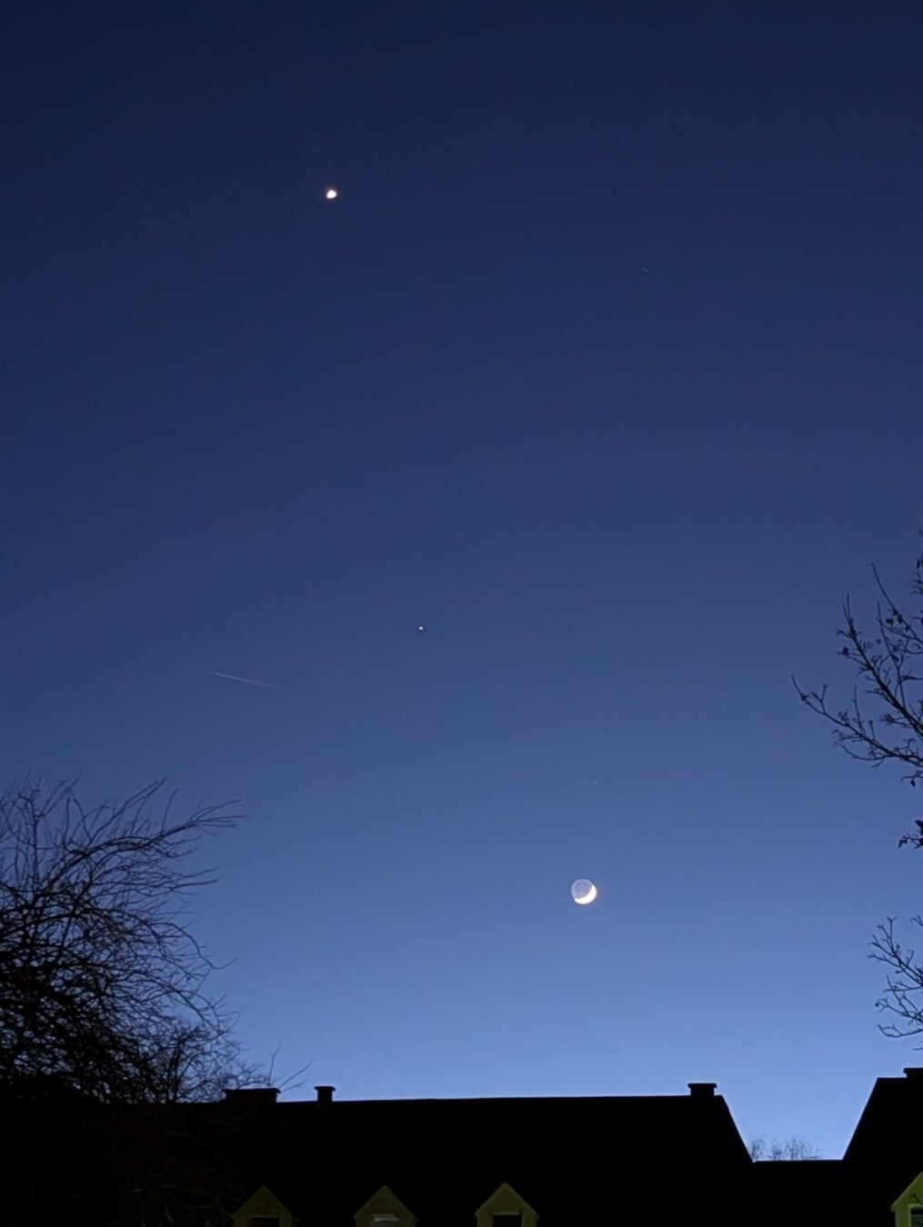 Blauer Abendhimmel über einem Stadtdach mit von links oben nach rechts unten: Venus als heller Fleck, Saturn als Punk und der Mond als grauer Kreis mit sehr schmaler Sichel.