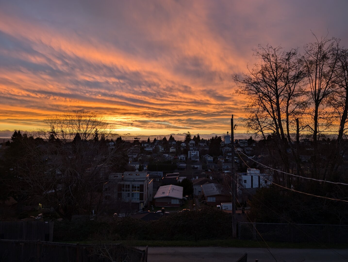 Picture looking over a neighborhood that covers a valley with the sun setting. The sky is covered in clouds but there a small band of clear where you can see sunlight.