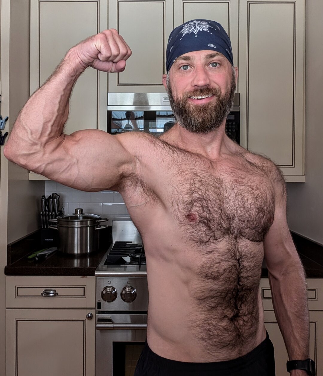 Bearded shirtless white guy wearing black shorts and a blue bandana flexes his right raised arm. The veins are STRONG this morning!