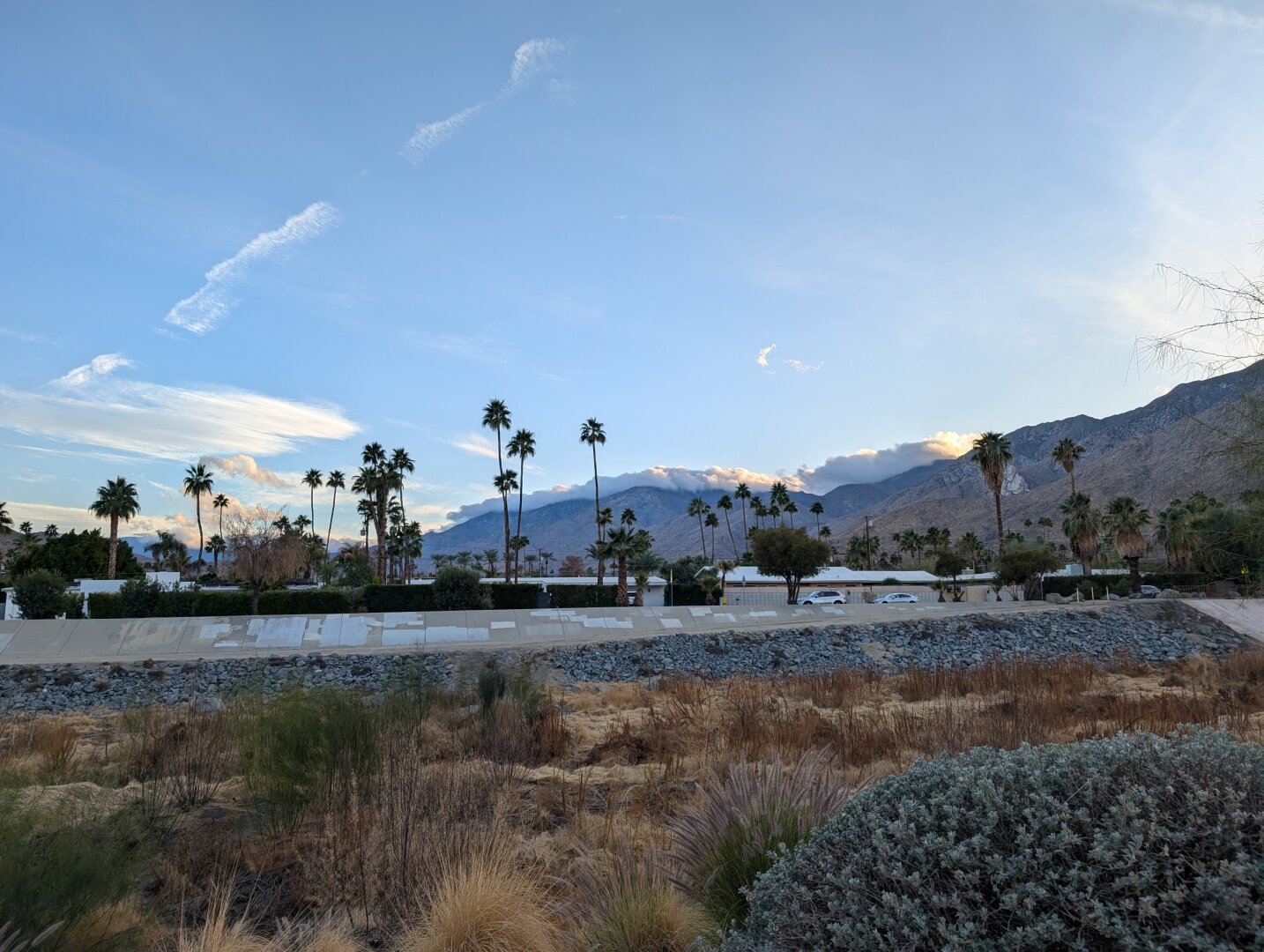 Photo across a dry flood channel with blue skies and mountains.