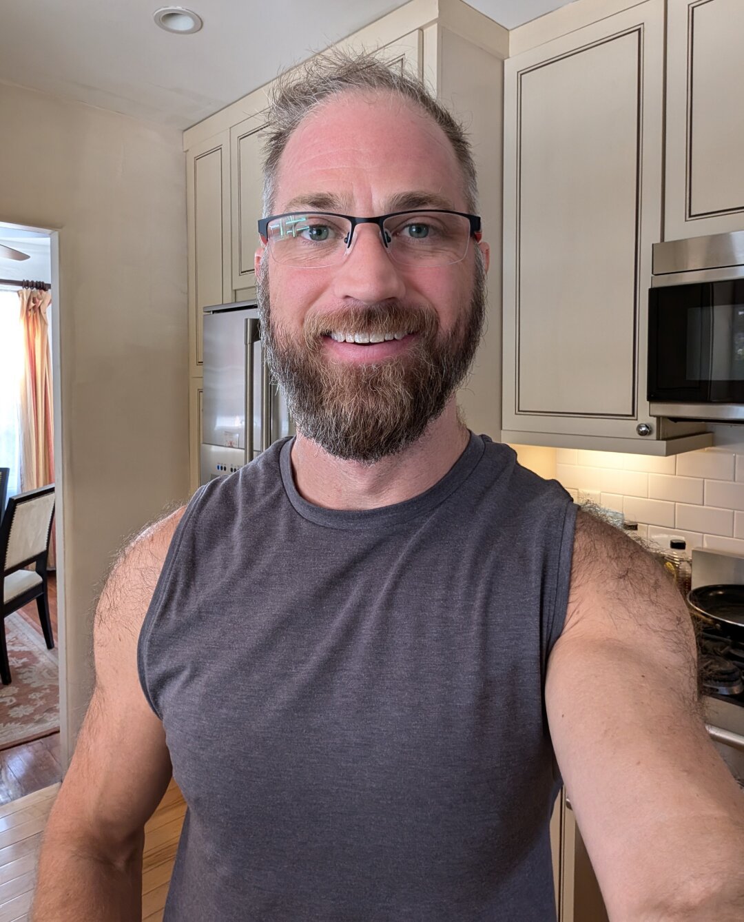 Bearded white guy with disheveled hair smiles for the camera. He's wearing glasses and a gray sleeveless shirt and standing in a kitchen.