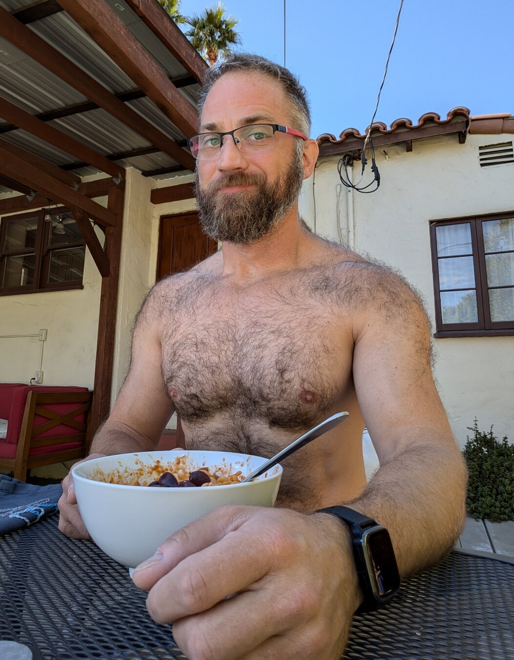 Shirtless, furry, bearded white guy sits on a table outside with a bowl of pasta with red sauce in front of him.