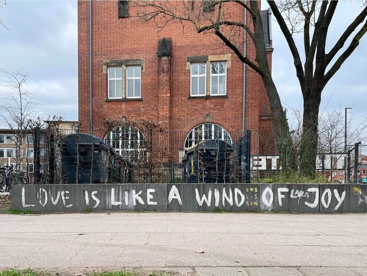 Gerollert in Weiß auf grauer Mauer: Love is like a wind of joy. (In Caps.) Hinter dem Mäuerchen städtische Kulisse mit Gebäuden, Zaun, Bäumen.