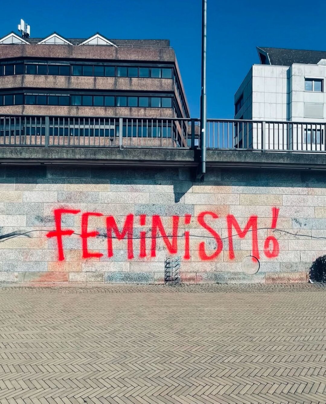 In Rot gesprüht (sehr groß) an eine Steinwand „feminism“ (in Caps). Davor Weg. Im Hintergrund darüber ein Geländer, Häuser, blauer Himmel.