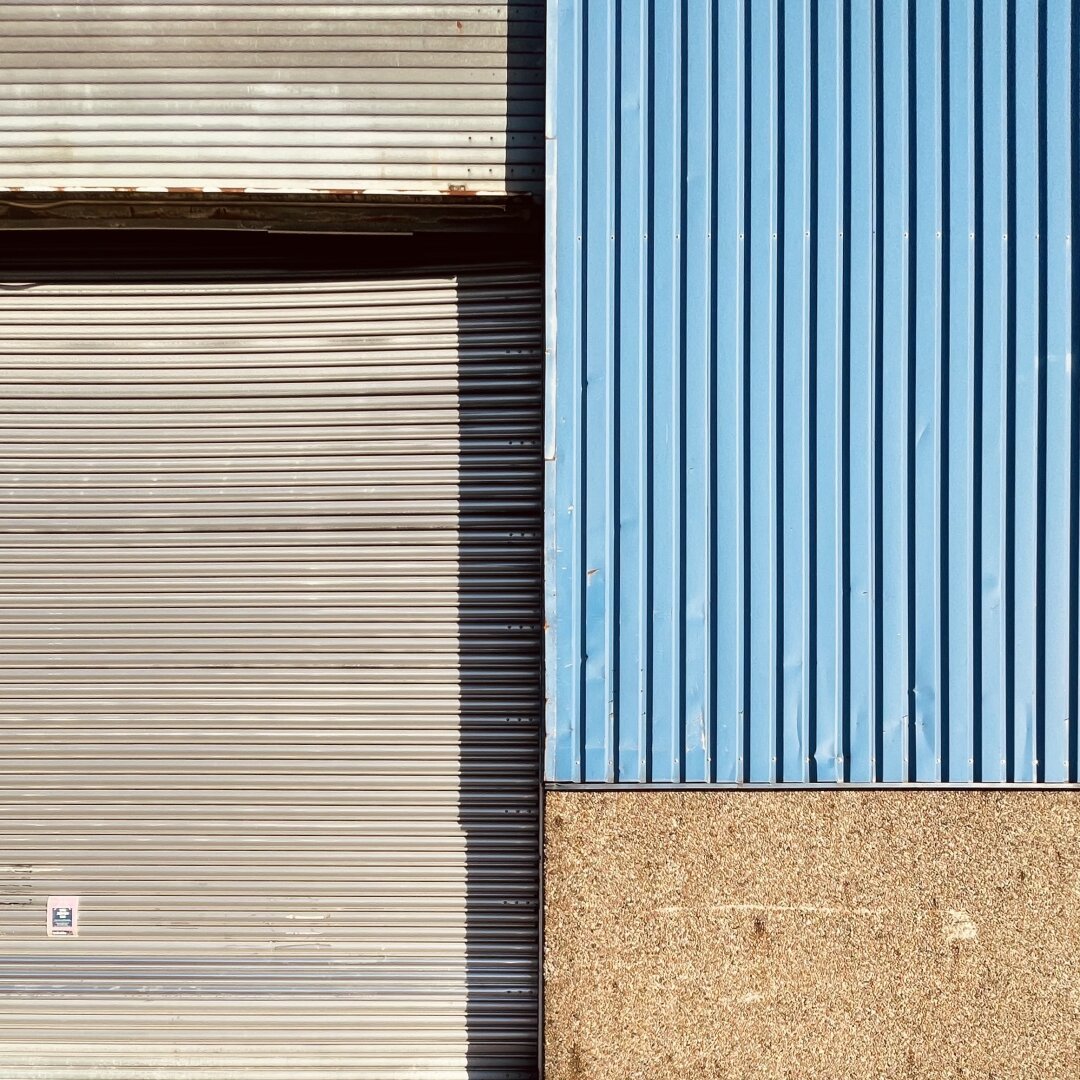 Der Ausschnitt einer Fassade einer Lagerhalle (oder ähnliches). Die linke Hälfte ist ein silberfarbenes Metalltor in Wellblechoptik im Anschnitt. Die rechte Hälfte ist Wand im Anschnitt. Der obere Teil ist blaues Wellblech, der untere Teil bräunlicher Waschbeton.