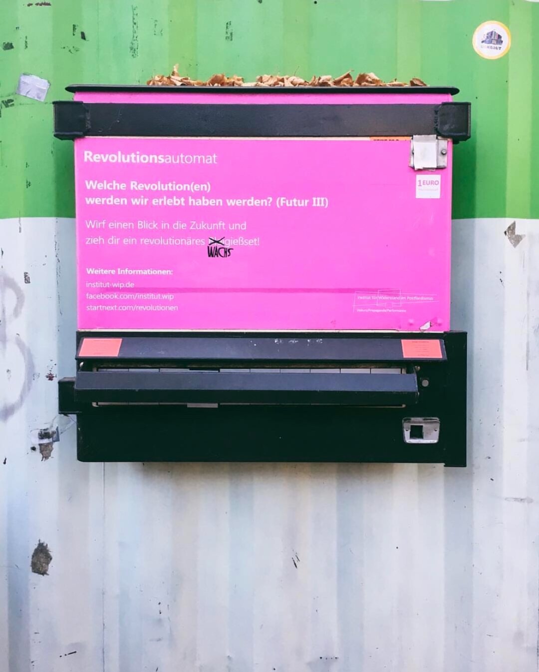 Ein umgewandelter Zigarettenautomat an einem weiß-grünen Container. Der Automat ist mit schwarzer und pinker Folie beklebt. Darauf folgender Text in Weiß:  Welche Revolution(en) werden wir erlebt haben werden? (Futur III) Wirf einen Blick in die Zukunft und zieh dir ein revolutionäres Wachsset. Weitere Informationen beim Institut WIP (facebook-Link) 1 EURO