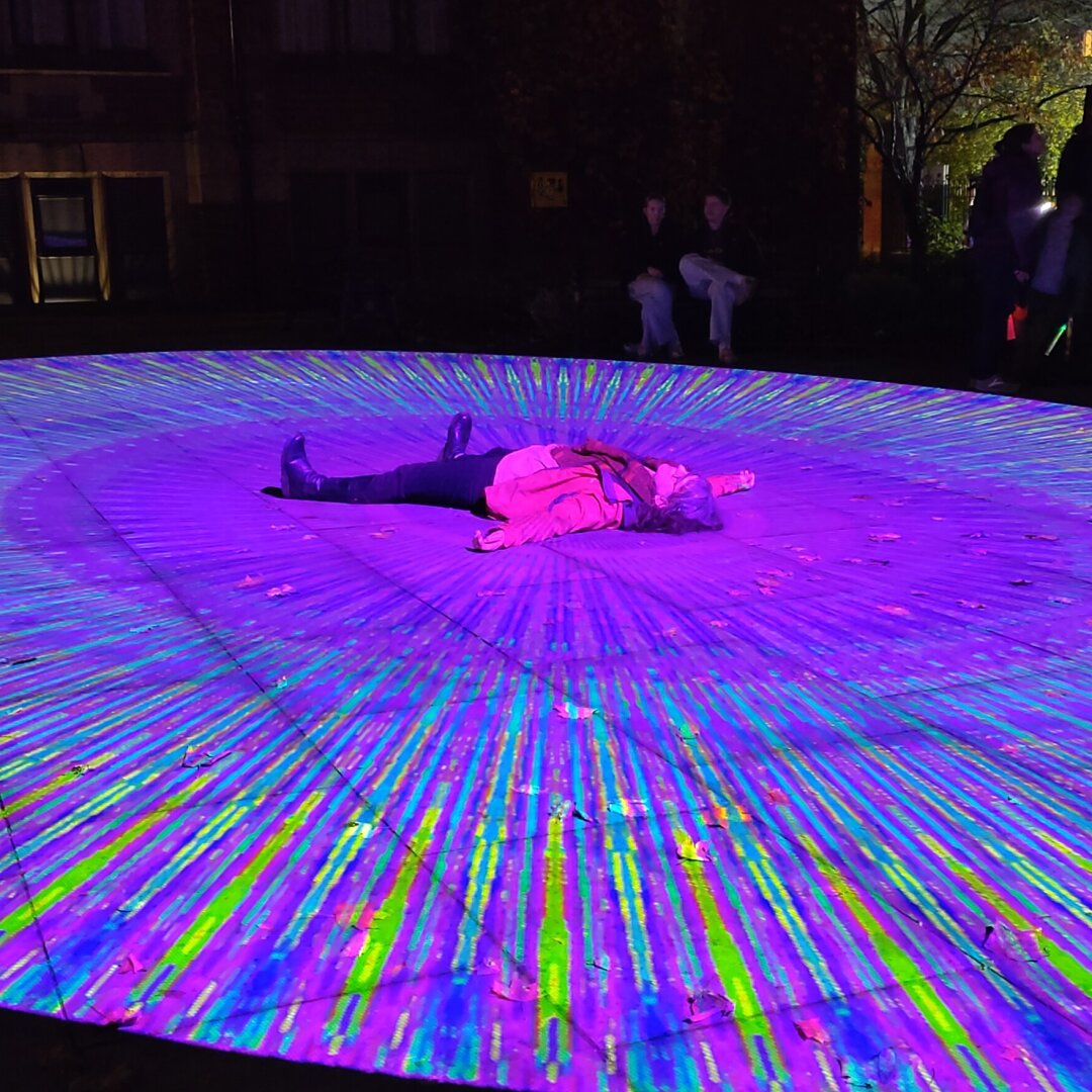 A person laid on their back on the ground - with their arms and legs spread out - in the middle of a large circular projection mapping. The projection is a repeating radial pattern of purples blues and greens perhaps every 5-10 degrees around the circle.