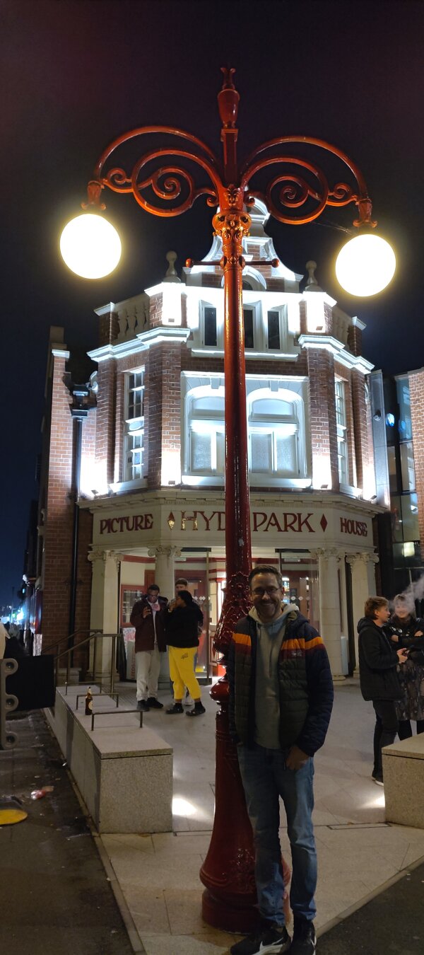 A man in a warm jacket stood next to an old fashioned lampost, at night, in front of an old cinema on a street corner.