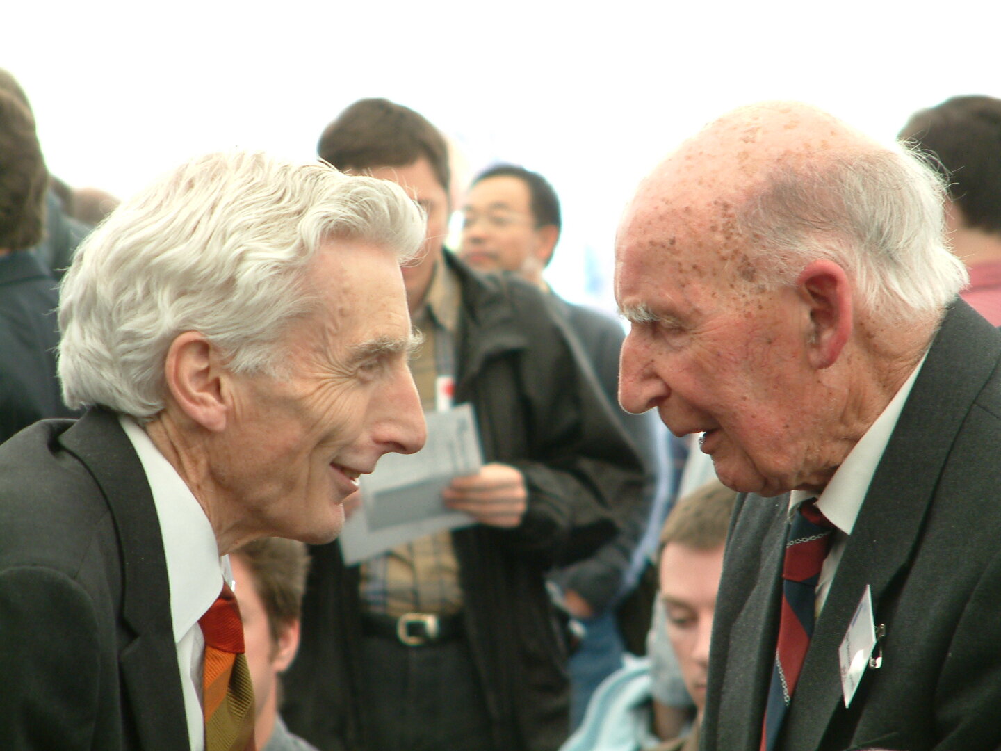 Two older white men facing each other. On the left is Martin Rees with white hair and a smiling expression. On the right is an elderly Bernard Lovell also with white hair but bald on top. Both are wearing dark suits with ties.