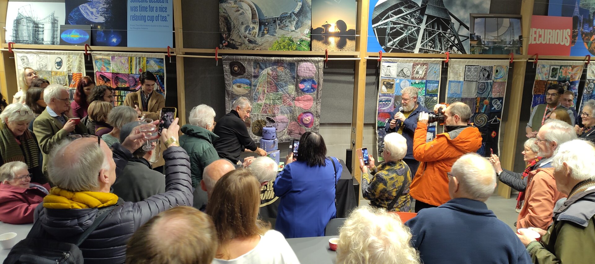 Director Mike Garret cutting a special, three-tiered lilac cake with a Lovell Telescope on top. In the foreground are lots of old Jodrellites. Behind are tapestries that are part of a special exhibition called "Codmic Threads".