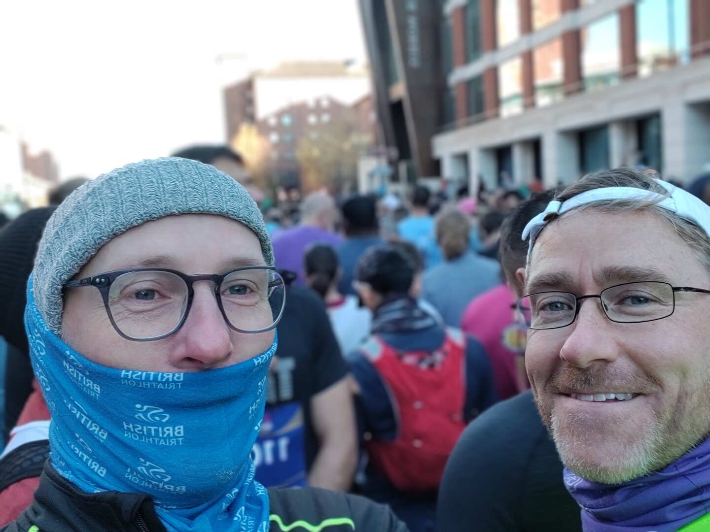 Two white men with glasses at the start of a running race with other runners behind. One has a woolly hat and buff up around the lower half of his face with a chilly nose. The other is smiling and not as cold.