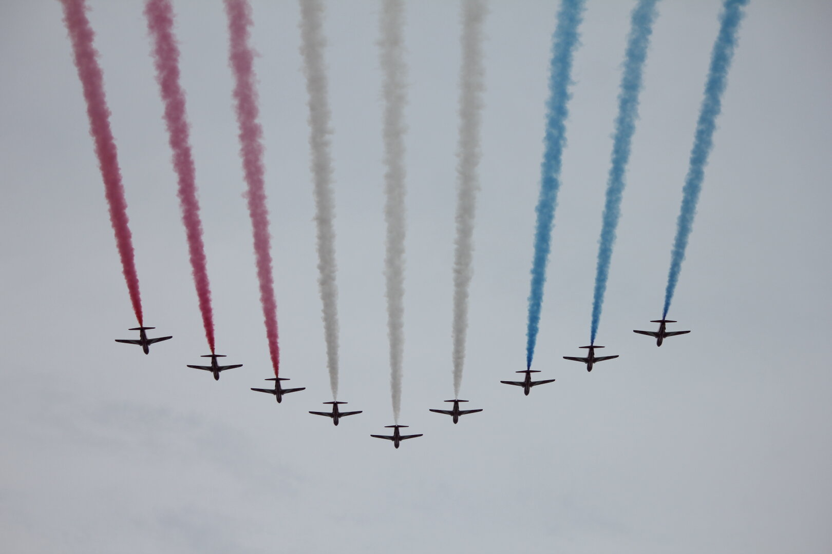 9 jets fly towards the bottom of the picture in a 'v' formation with red, white, and blue smoke trails behind.