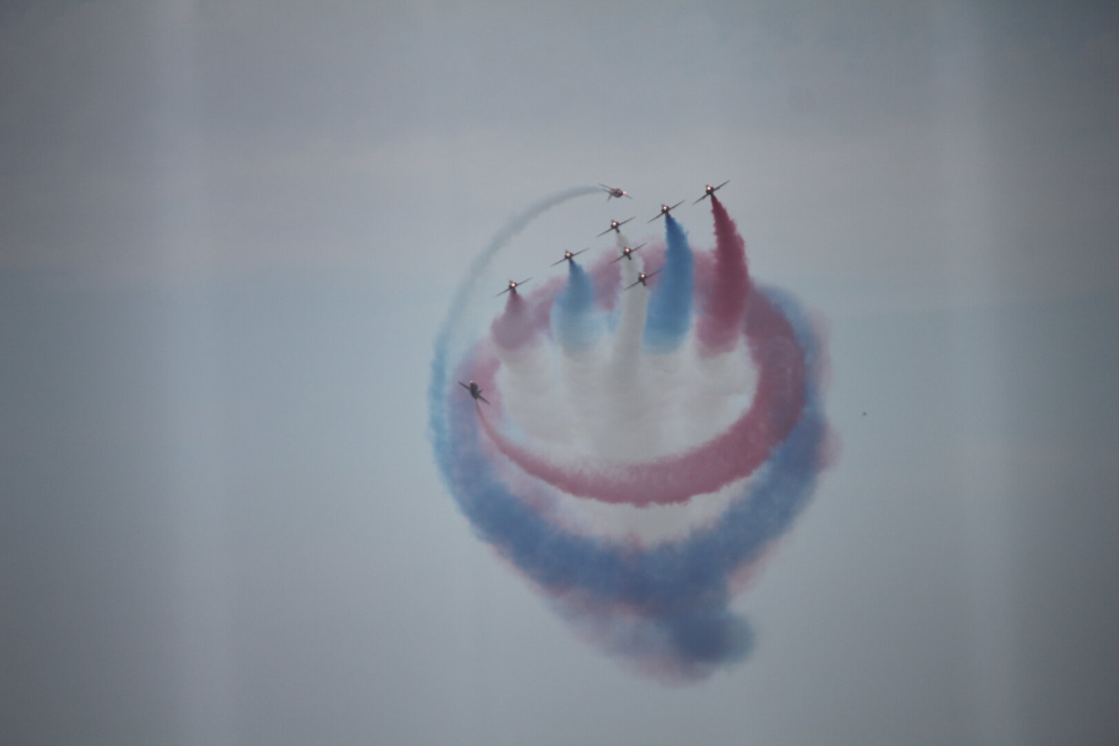 A formation of 7 Red Arrows make an arrow flying towars the camera whilst two Red Arrows circle them with red and blue smoke trails.