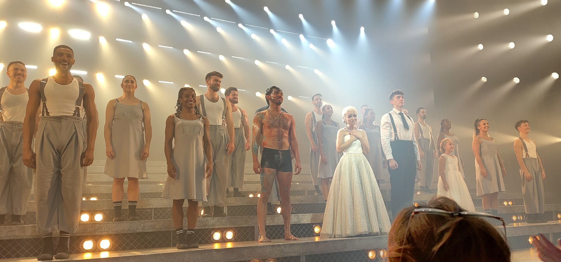 The cast of Evita stood on a stepped stage at the end of the show. They are wearing white vests with either grey high-waisted trousers or grey knee length dresses. The three main characters (and a young girl) are stood at centre stage. Che is on the left just wearing black boxers and is dripping in red/blue/white paint. Juan is stood right to the right of centre with black trousers, white shirt, braces, and black tie. Rachel Zegler as Eva is in the middle wearing a long white dress and clutching her hand to her chest. There are many spotlights in the background all pointing towards Eva.