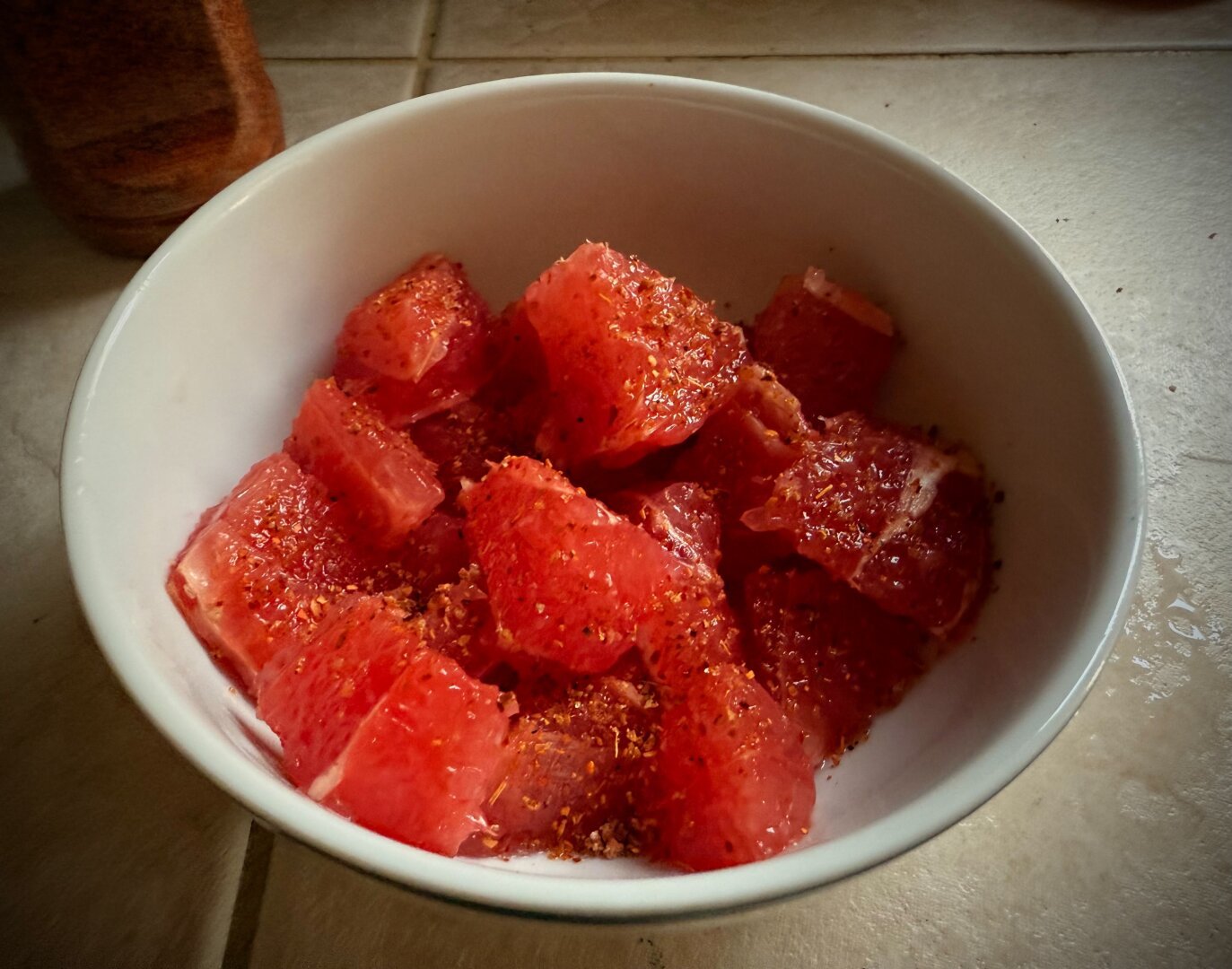 A white cereal bowl mostly full of ruby red grapefruit with chili seasoning sprinkled on top