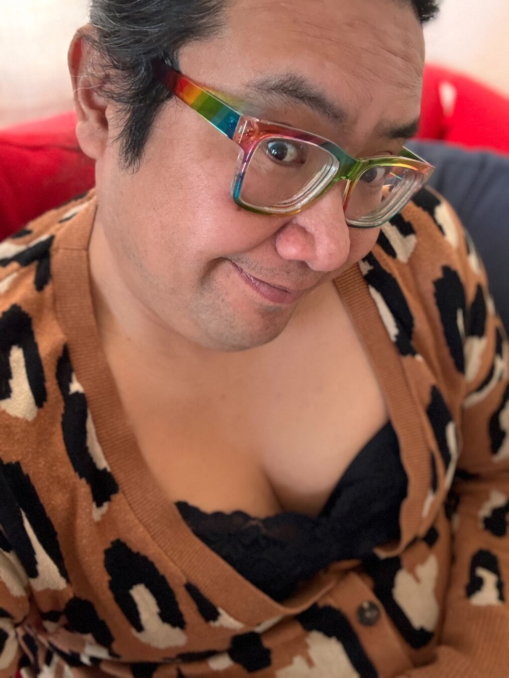 Selfie of a tan person wearing a lacy black camisole and a leopard print cardigan, they are showing a bit of cleavage and have a slight beard shadow