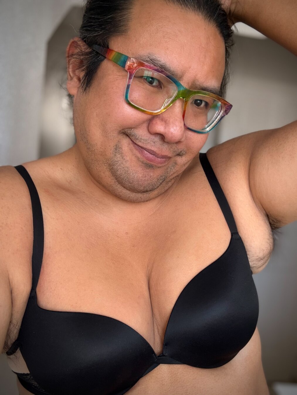 Selfie of a tanned person with rainbow glasses wearing a black bra, they have a five o'clock shadow