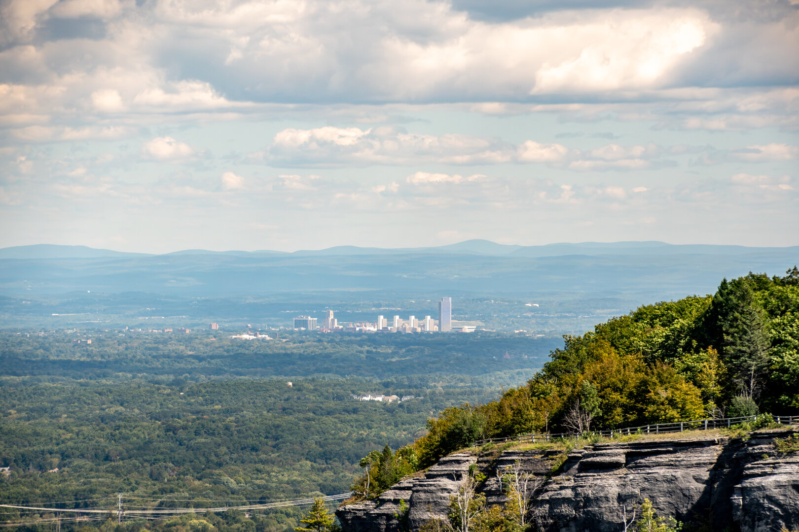A skyline of a small city surrounded by green mountains, puffy clouds in the sky and closer cliff with trees in the bottom corner