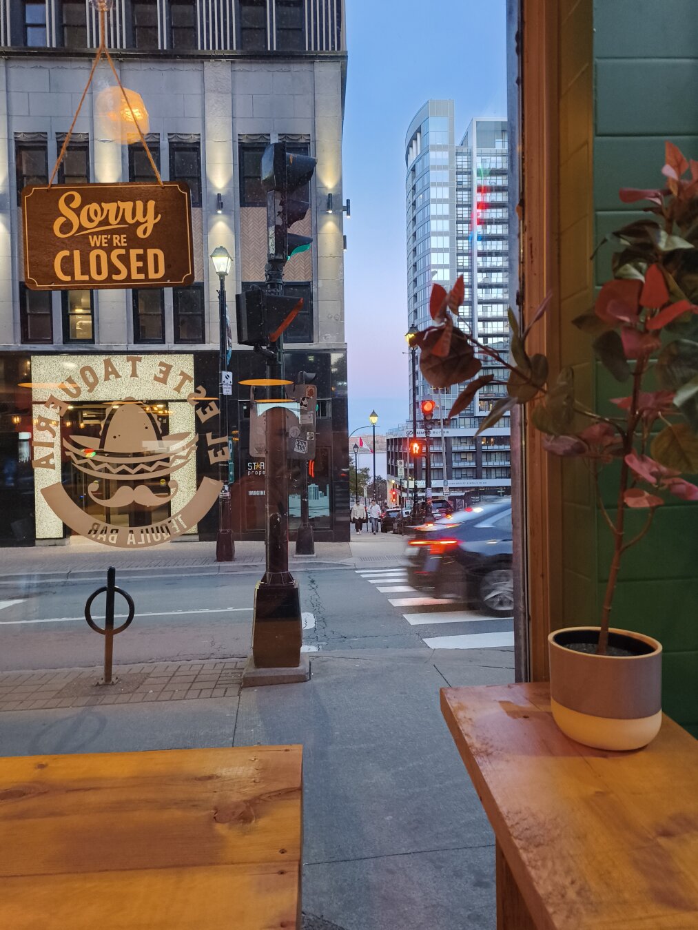 Showinf a city scene, there is a large highrise in the distance, a car zooming by a restaurant window. You can see another large building across the street that has uplights on it. There is an open/closed sign on the window, and tou can see wooden benches lined up against the inside of the restaurants windows. There is a small potted plant on one of the benches.