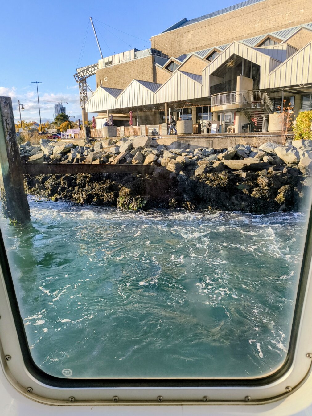 Showing the view from a ferry window, it's rectangular with rounded corners. You can see the swirling turquoise water being disturbed by the boat. Nearby behind that, there is a dark rocky shore and a large ferry terminal building with multiple pointed roofs forming an overhang. The bright blue sky is behind that.