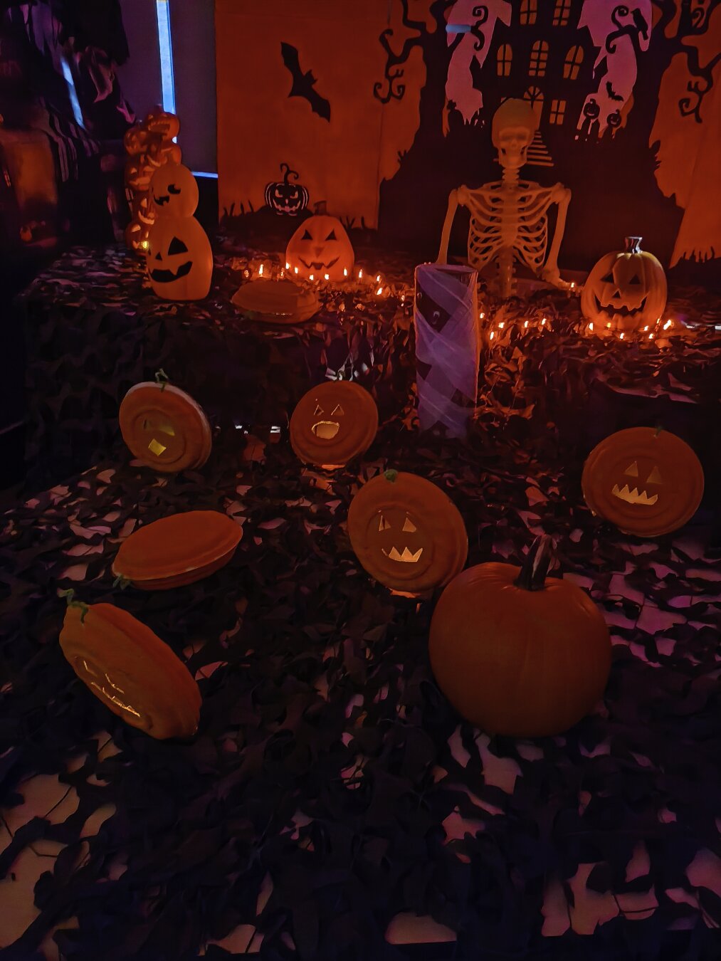 Showing a scene with paper pumpkins with carved faces, with a backdrop of a spooky house with Gothic looking trees and windows. There are orange lights and a mesh like black cloth covering white sheet where the pumpkins are placed on. There is also a plastic skeleton sitting behind the pumpkins.