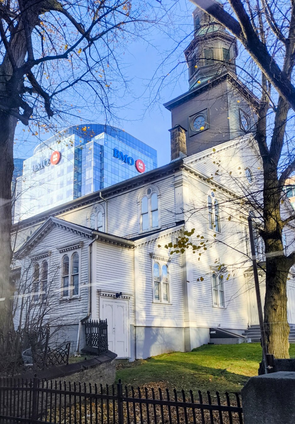 Showing a white wooden church from a corner on angle. Behind looming over it is a cube like blue glass building with the BMO logo on it. The sky is blue. There is some green grass in front of the church, there is reflected light shining on the church from the otherside of the road from a large skyscraper.