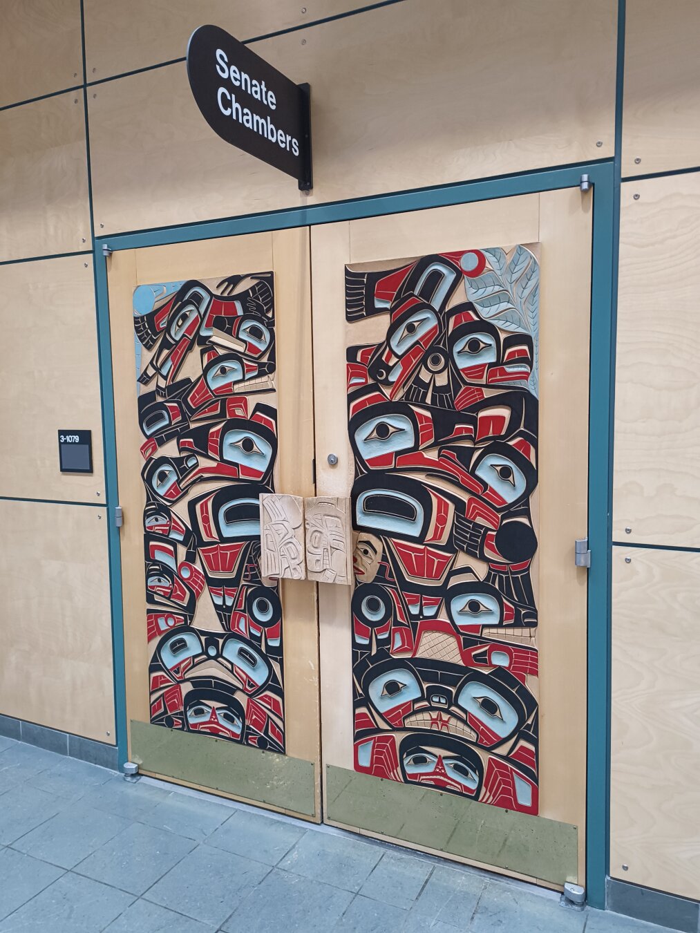 Showing a wooden door carved with aboriginal artwork in the style of West coast BC nations. It's colorful with red and blue and black and pine wood.