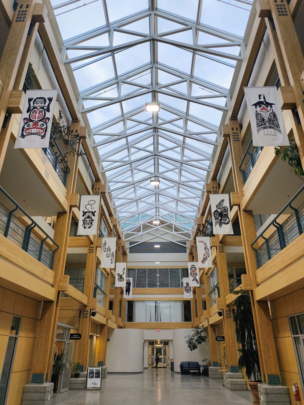 Showing a glass topped atrium with wooden beams and pillars on each side. There is aboriginal artwork hung on each pillar and there is light coming through the atrium lighting it up.