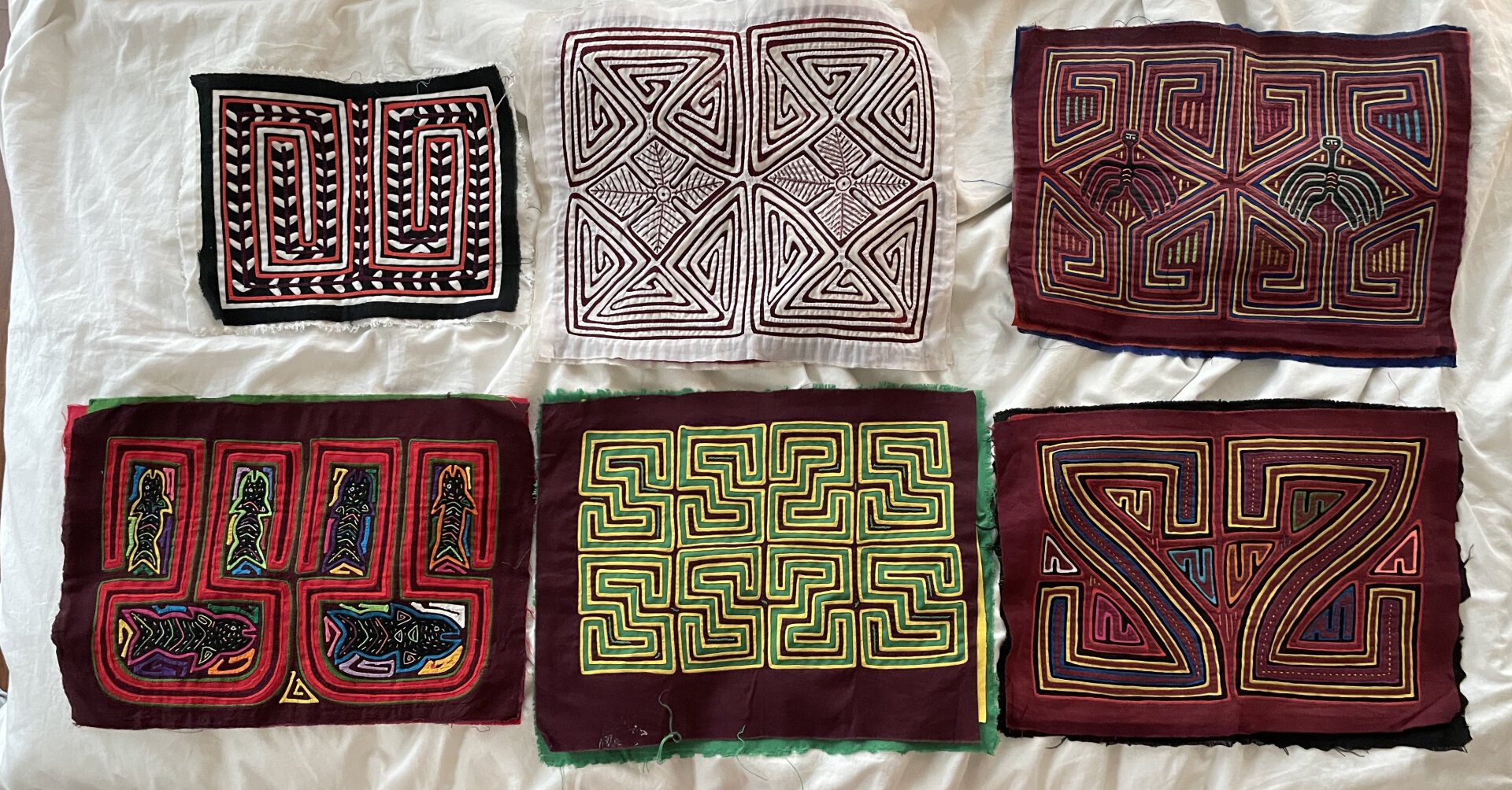 6 molas laid out on a bed. They all have red and are geographic patterns made by layers of fabric with holes cut into them and then appliqued on top. There are typically three layers of fabric.