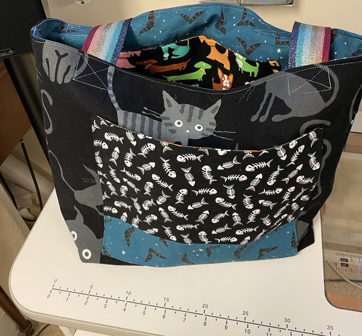 Tote bag made from black Japanese fabric with grey electrified kitties.  There’s a large pocket with fish skeletons and bats on blue. A kitty face is poking just above the pocket.  Inside you can see a blue fabric with bats and an interior pocket with colorful cartoon dogs.  The straps are a sparkly rainbow.