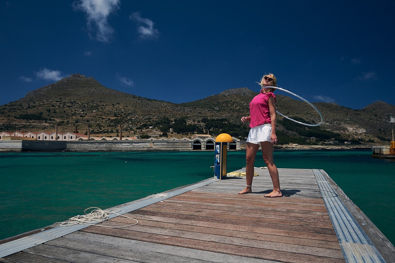 A woman is standing on a pier within a small harbour, she has a hula hoop circling around her neck. The seawater inside the harbour is turquoise with blue patches. At the end of the harbour the ruins of an old tuna factory are seen. Behind the ruins small mountains or hills are seen with the ruins of a castle on top of the largest one. Above a dark blue sky with only patches of white clouds are seen.