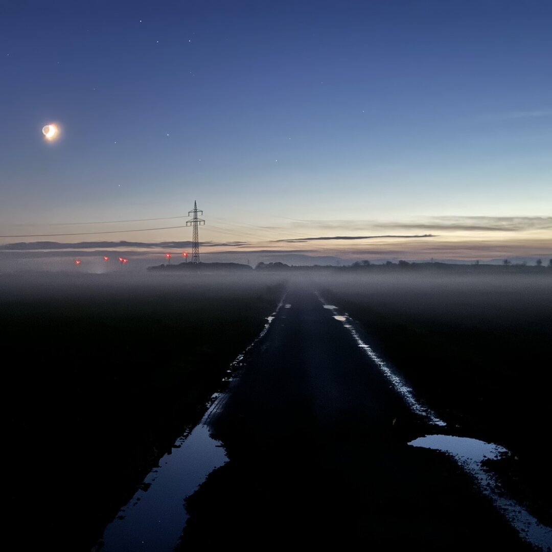 The lower part of the photo shows a road into a field. It’s very dark, nearly black, but there are puddles of water on the road which reflect the blue sky. Towards the horizon it’s getting quite misty, though some trees, a power pole and the red warning lights of wind power plants are visible. The upper side displays a blue sky with a bright moon and some stars.