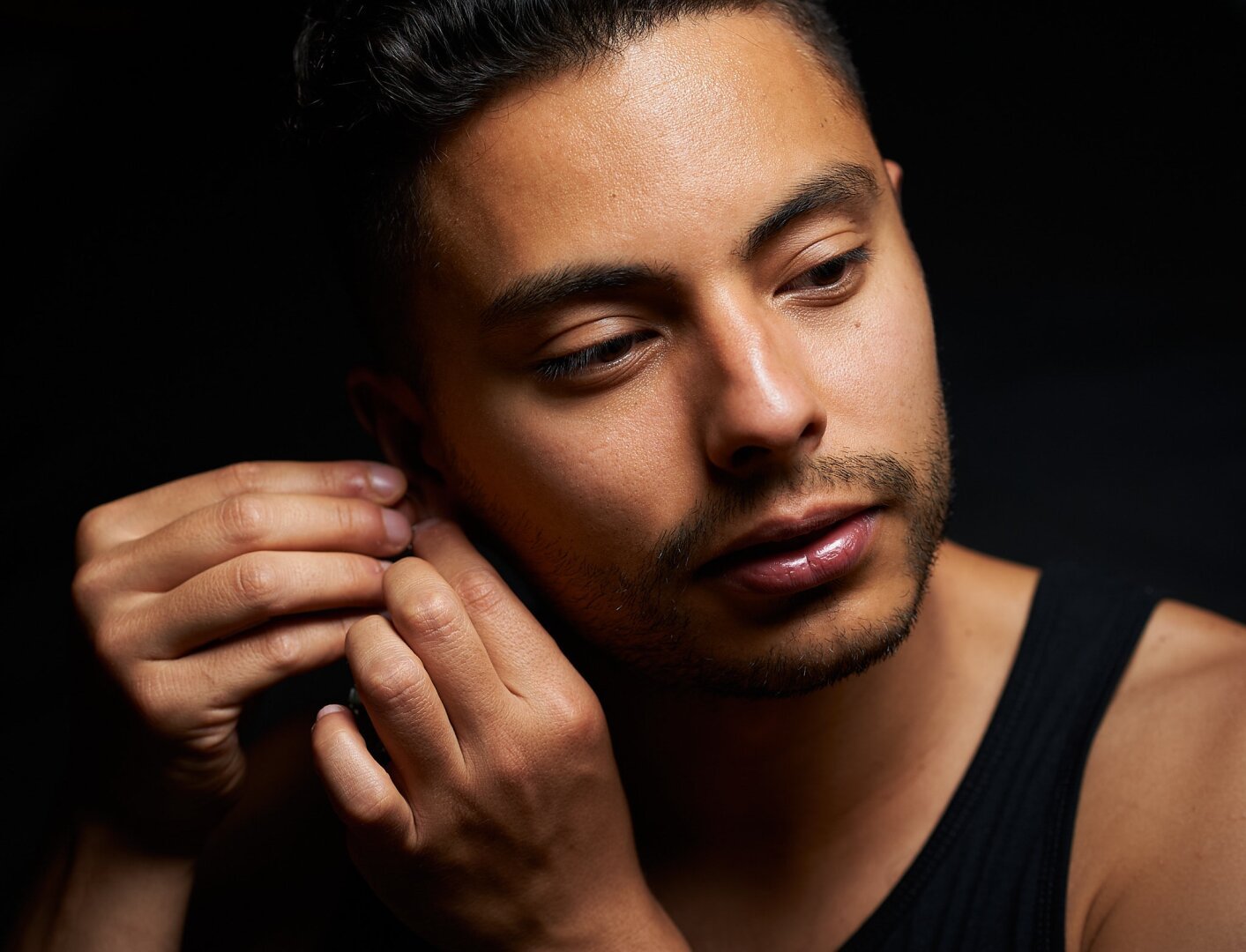 The photo shows the portrait of a man who is adjusting an earring with both hands on his right earlobe. His face, shoulders and hands are visible in front of a dark background.