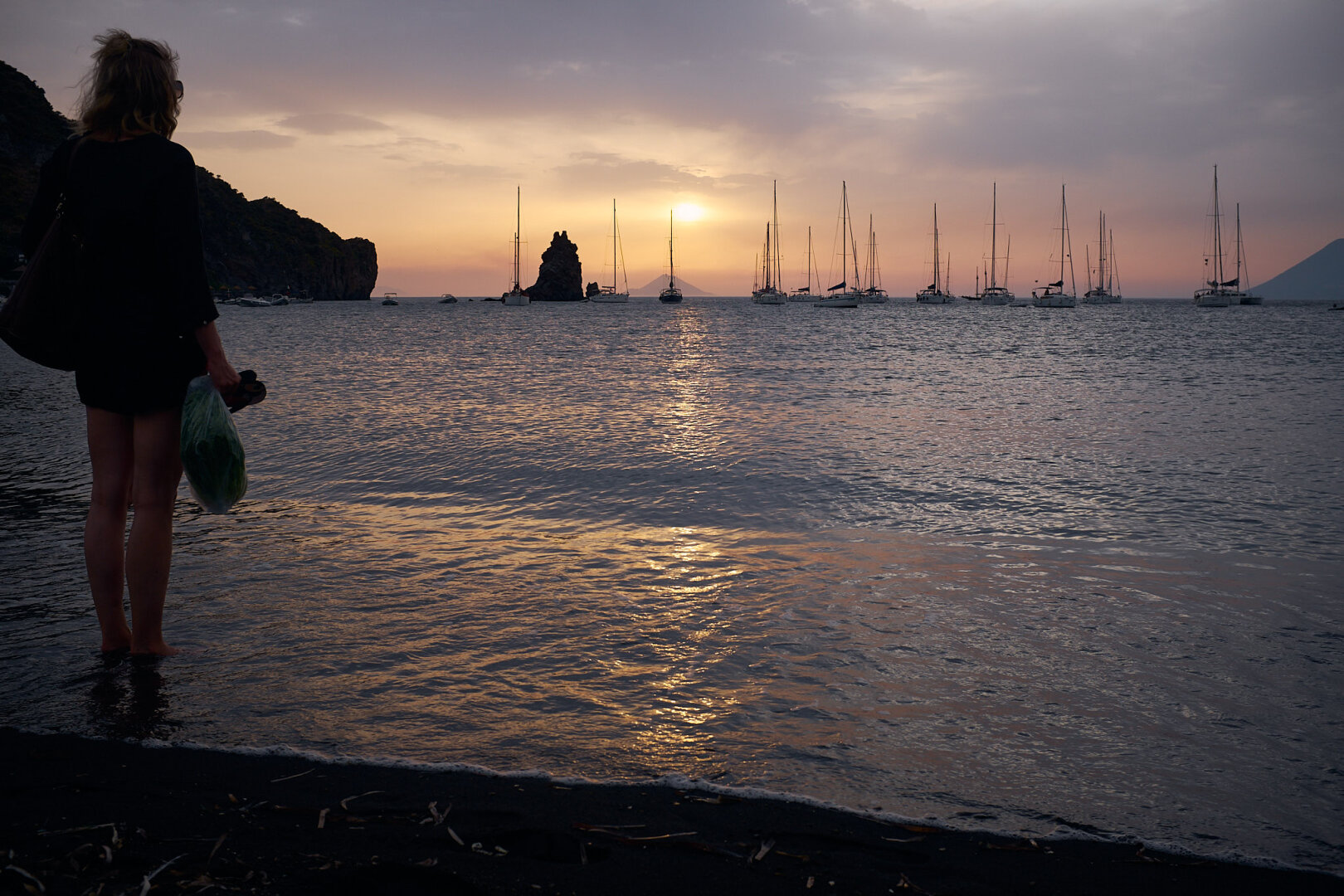 The photo shows a bay in the Mediterranean Sea with sailing boats anchored for the night. Behind the sailing boats the sun is setting. The sky is cloudy, but the sun is still visible and the orange colors are reflected by the sea. At the beach to the left the silhouette of a woman waiting to be picked up and brought to one of the sailing boats is visible.