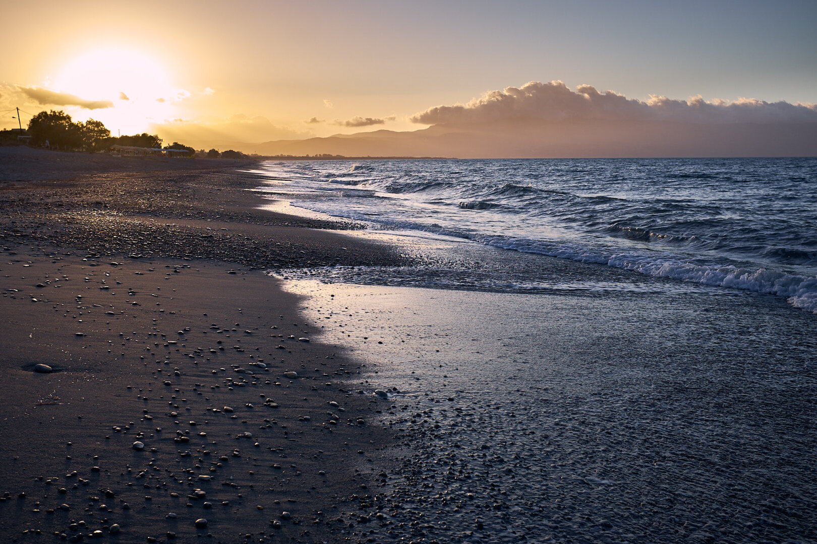 The photo shows a beach on the left side with the sea on the right side during sunset. The sun is setting on the left upper side over land. The sea, the wet part of the sand and the stones on the dark appearing sand are reflecting the golden light.