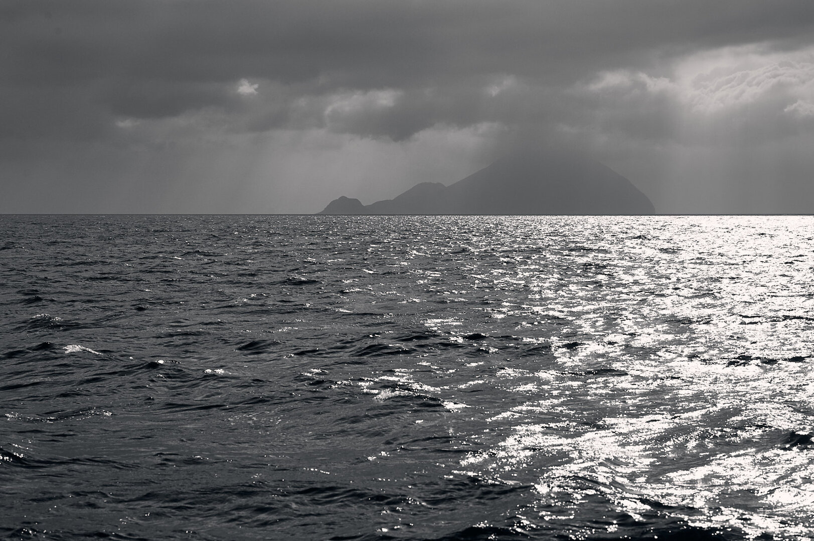 The photo shows the calm Mediterranean Sea in the lower two-thirds, reflecting the harsh sunlight. In the background the island of Filicudi is visible with clouds and rain. The photo is black and white and has a high contrast.