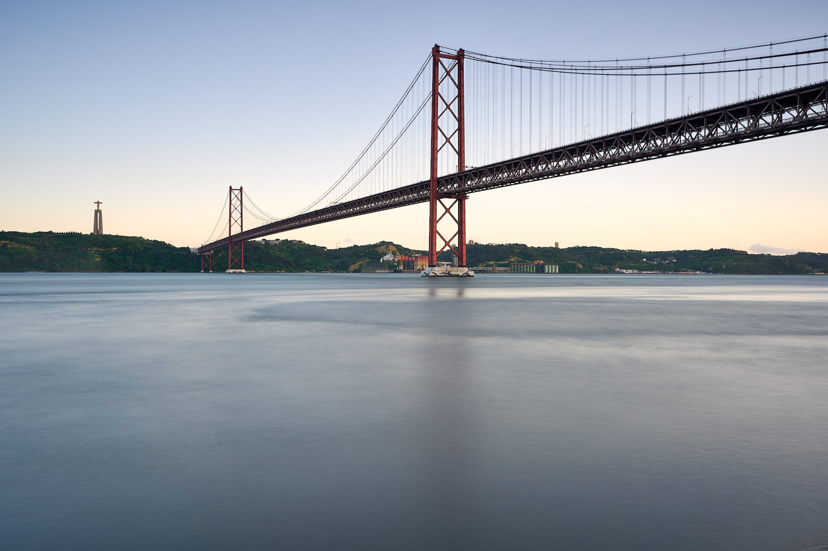 The photo shows a long exposure of the Tejo river in the foreground with the red bridge ‚25 de Abril‘ coming in from the upper right towards the upper left where it connects the municipality of Almada with Lisbon city center. On the upper left the statue ‚Sanctuary of Christ the King‘ is visible in the far. The blue sky with a golden line is visible behind the bridge.