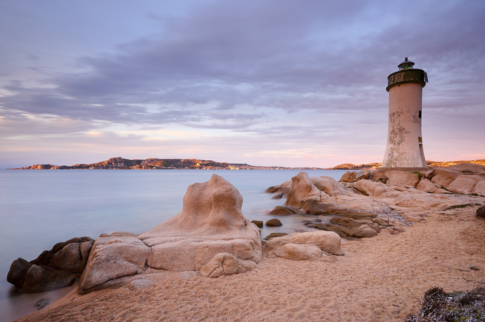 The photo shows the beach with some rock formations in front of the milky turquoise sea. To the right a lighthouse reaches up into the blue, slightly violet and orange sky. There are some clouds in the sky. In the back the island of ‚La Maddalena‘ is enlightened by the last rays of the setting sun.