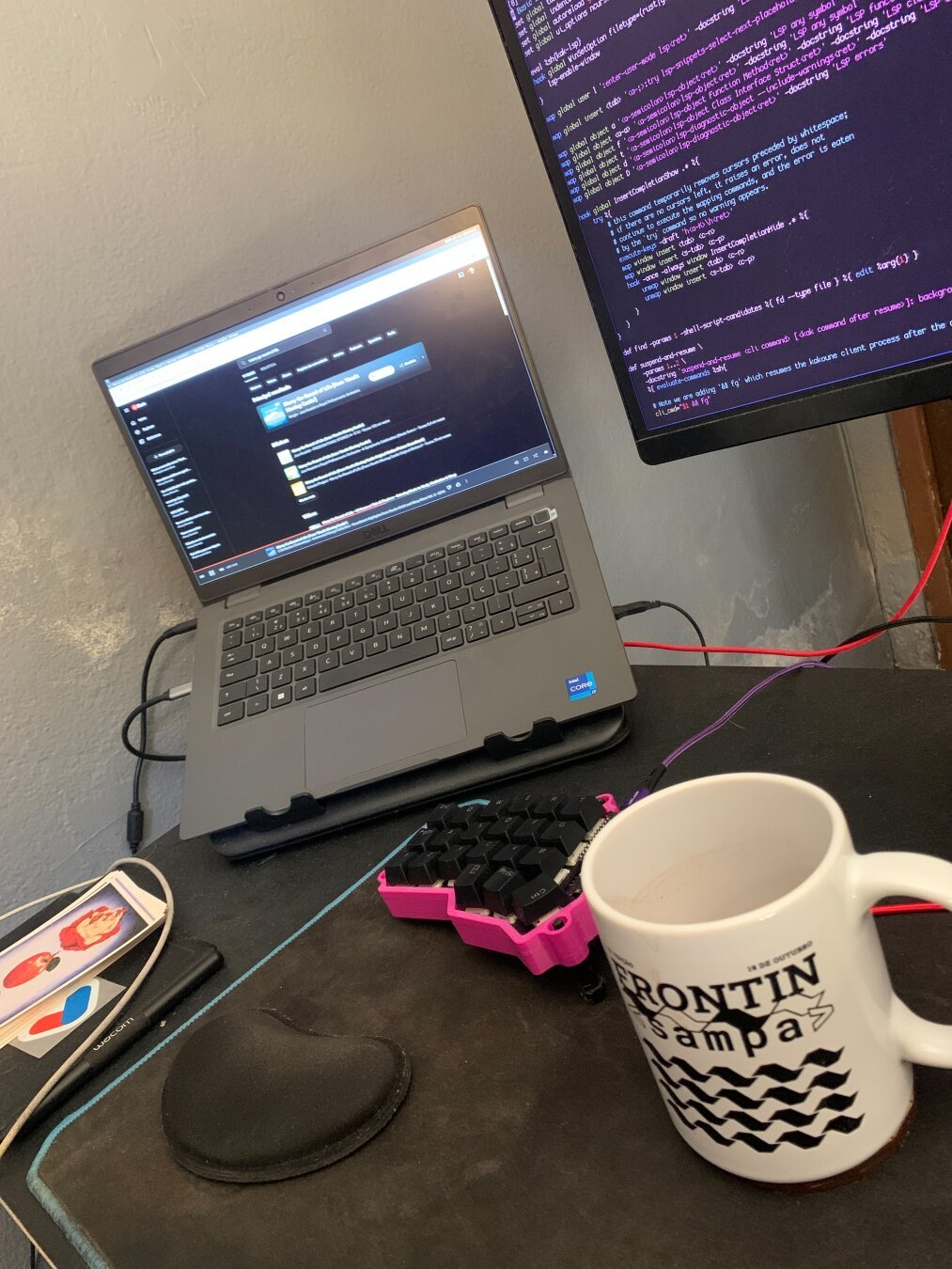 Uma foto com um notebook , metade de um teclado ergonomico rosa, um monitor suspenso por braço mecânico e uma caneca escrito Frontin sampa