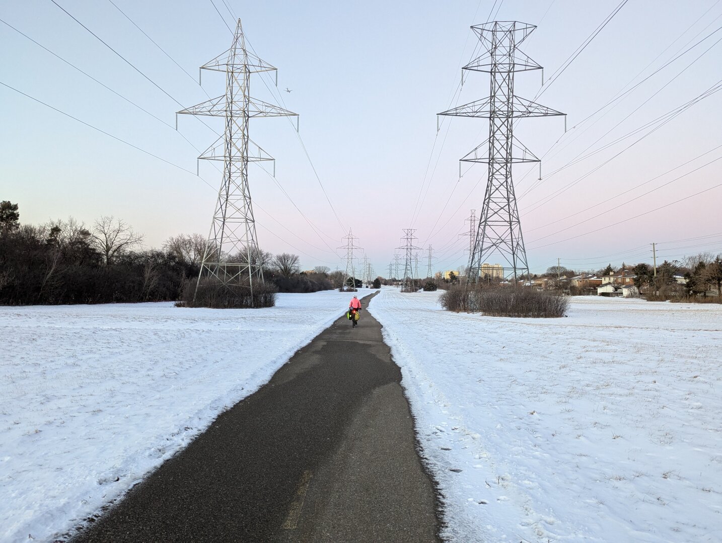 A long view of a paved path leading into the distance through a field covered in fresh snow. A cyclist in a pink jacket is seen further down the path. Large metal hydro towers with connecting power lines punctuate the landscape. The sky is a pale blue with hints of pink from the rising sun.